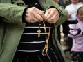 Dłonie starszej osoby trzymające różaniec. W tle widać inne osoby, w tym dziecko. To modlitwa o pracę dla syna.