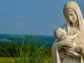 Kamienna Matka Boża ostrzega, tuląc Dzieciątko Jezus. W tle zielone wzgórza i błękitne niebo.