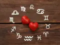 Dwa czerwone serca otoczone symbolami zodiaku, w tym znaki Wagi i Bliźniąt, na drewnianym tle.
