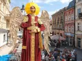 Gigantyczny Archanioł w czerwonej szacie z aureolą i złotymi skrzydłami, jakby wznosił modlitwę do archanioła. Ludzie spacerują ulicą.