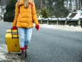 Kobieta w żółtej kurtce i czerwonych rękawiczkach ciągnie żółtą walizkę. W tle zimowy krajobraz. Modlitwa o szczęśliwą podróż.