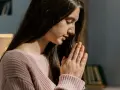 Młoda kobieta w różowym swetrze, z dłońmi złożonymi do modlitwy. Może to być modlitwa o pieniądze, o zdrowie lub o spokój.
