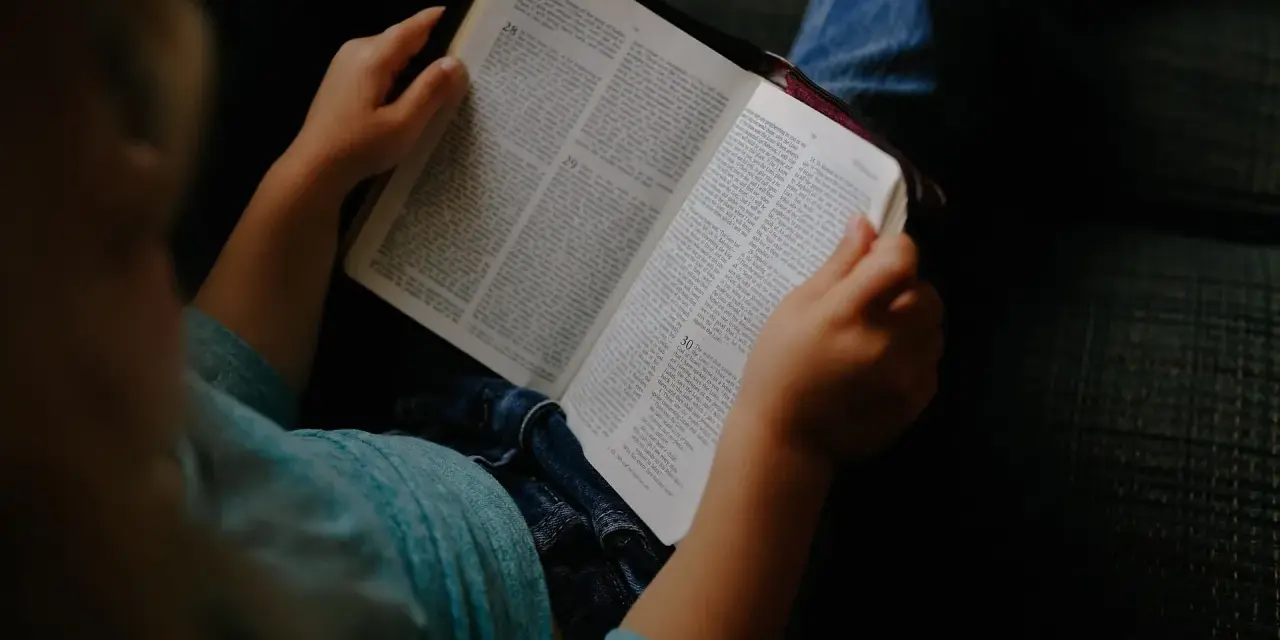 Dziecko w niebieskiej koszulce trzyma otwartą Biblię, przygotowując się do modlitwy przed czytaniem Pisma Świętego.