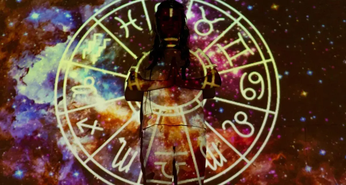 Postać w centrum, otoczona kosmiczną mgławicą, z projekcją ogniste znaki zodiaku na ciele.