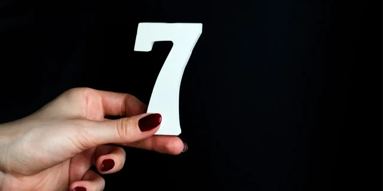 Dłoń z ciemnoczerwonym lakierem trzyma białą cyfrę 7. To symbol liczby duszy 7, oznaczającej mądrość i intuicję.