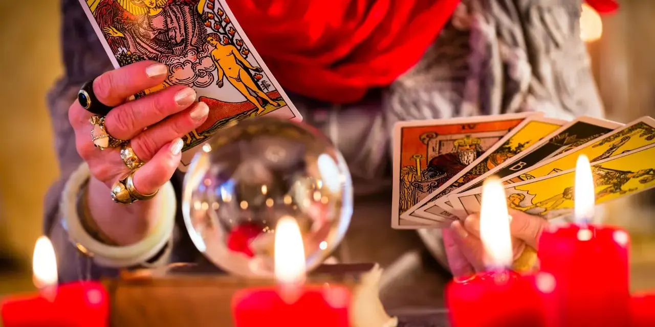 Dłoń z pierścieniami trzyma karty tarota, przygotowując tarot tygodniowy dla Bliźniąt. W tle kryształowa kula i czerwone świece.