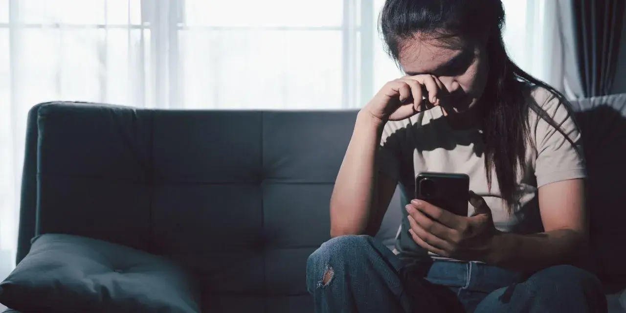 Zmartwiona kobieta siedzi na kanapie, trzymając telefon. Czyta wiadomości, które mogą dotyczyć jej związku z rakiem i wagą.
