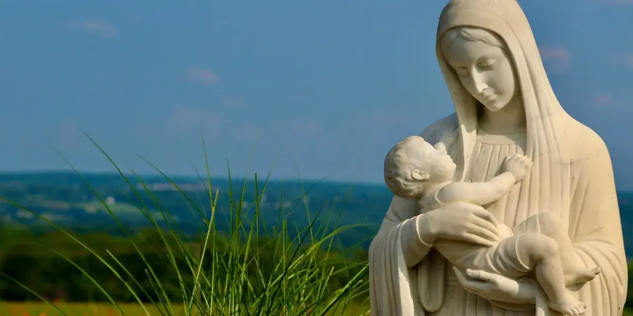 Kamienna Matka Boża ostrzega, tuląc Dzieciątko Jezus. W tle zielone wzgórza i błękitne niebo.