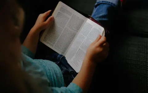 Dziecko w niebieskiej koszulce trzyma otwartą Biblię, przygotowując się do modlitwy przed czytaniem Pisma Świętego.