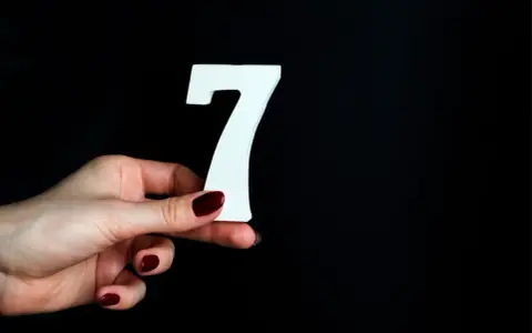 Dłoń z ciemnoczerwonym lakierem trzyma białą cyfrę 7. To symbol liczby duszy 7, oznaczającej mądrość i intuicję.