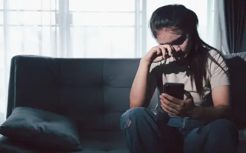 Zmartwiona kobieta siedzi na kanapie, trzymając telefon. Czyta wiadomości, które mogą dotyczyć jej związku z rakiem i wagą.