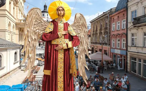Gigantyczny Archanioł w czerwonej szacie z aureolą i złotymi skrzydłami, jakby wznosił modlitwę do archanioła. Ludzie spacerują ulicą.