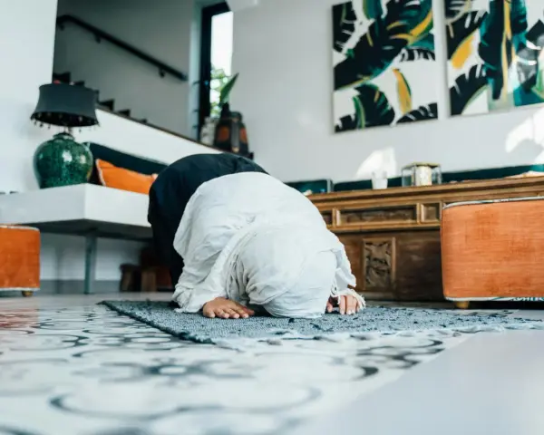 Kobieta w hidżabie klęczy na dywaniku, wykonując modlitwy o pokój. W tle widać nowoczesne wnętrze z dziełami sztuki.