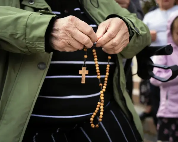 Dłonie starszej osoby trzymające różaniec. W tle widać inne osoby, w tym dziecko. To modlitwa o pracę dla syna.