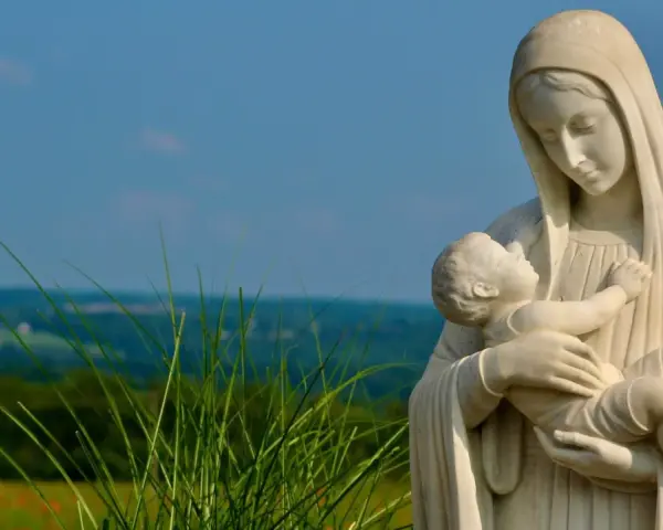 Kamienna Matka Boża ostrzega, tuląc Dzieciątko Jezus. W tle zielone wzgórza i błękitne niebo.