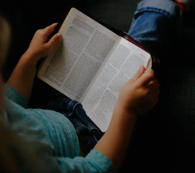 Dziecko w niebieskiej koszulce trzyma otwartą Biblię, przygotowując się do modlitwy przed czytaniem Pisma Świętego.