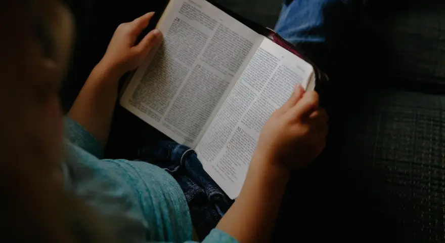 Dziecko w niebieskiej koszulce trzyma otwartą Biblię, przygotowując się do modlitwy przed czytaniem Pisma Świętego.