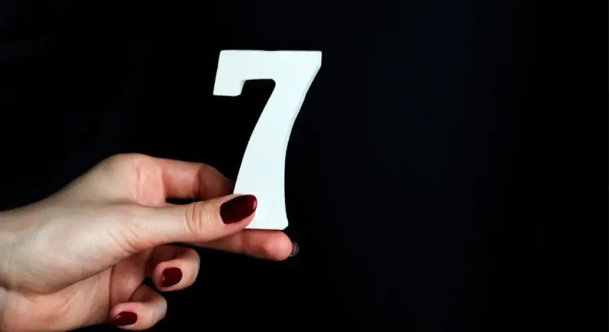 Dłoń z ciemnoczerwonym lakierem trzyma białą cyfrę 7. To symbol liczby duszy 7, oznaczającej mądrość i intuicję.