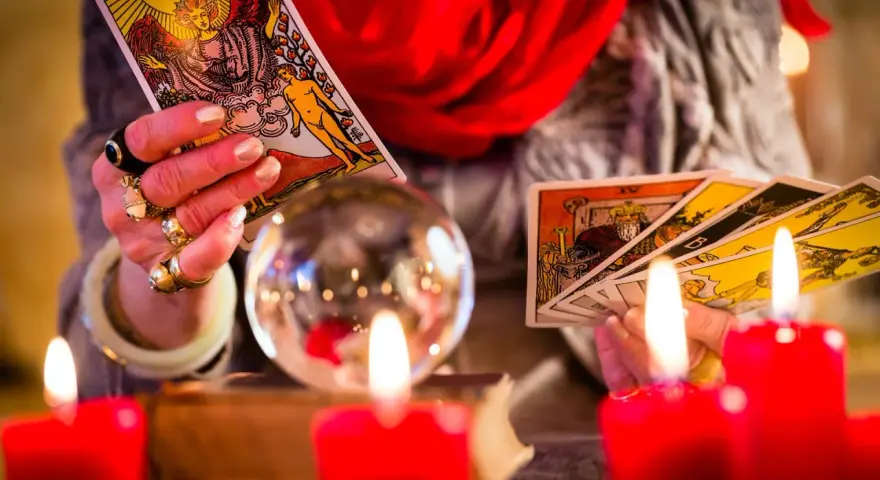 Dłoń z pierścieniami trzyma karty tarota, przygotowując tarot tygodniowy dla Bliźniąt. W tle kryształowa kula i czerwone świece.