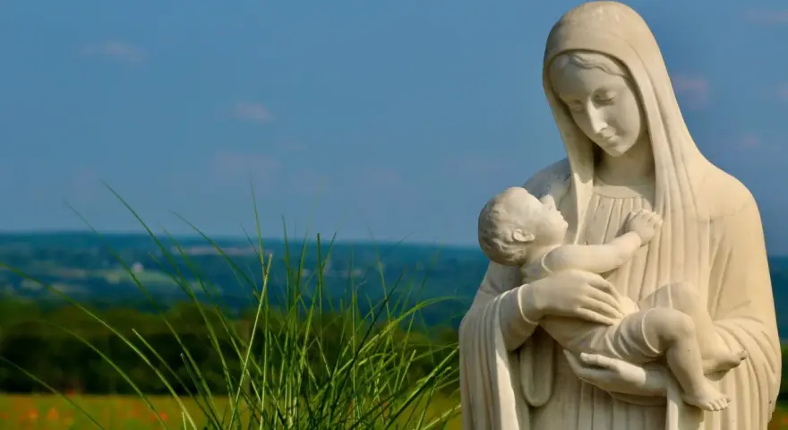 Kamienna Matka Boża ostrzega, tuląc Dzieciątko Jezus. W tle zielone wzgórza i błękitne niebo.