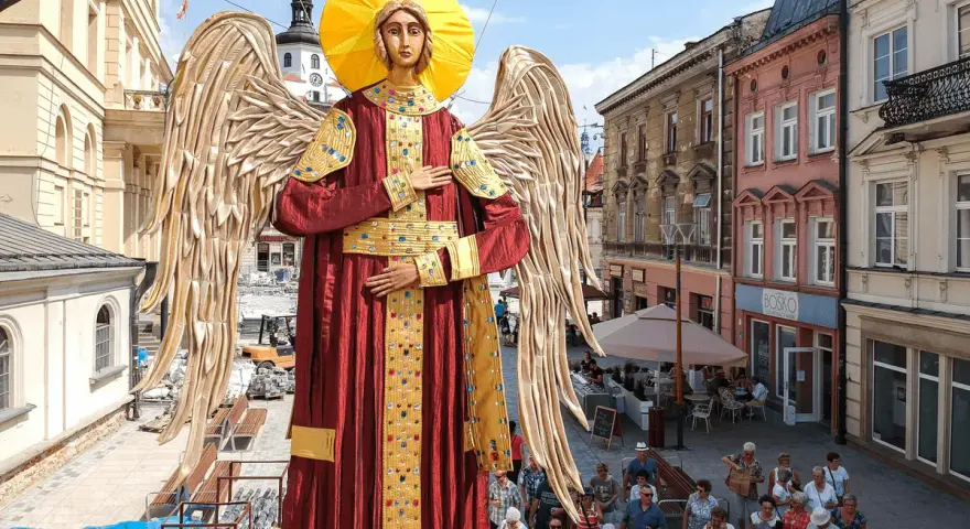 Gigantyczny Archanioł w czerwonej szacie z aureolą i złotymi skrzydłami, jakby wznosił modlitwę do archanioła. Ludzie spacerują ulicą.