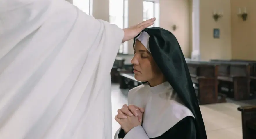 Kapłan błogosławi siostrę zakonną w kościele. W tle widać ławki i okna. To piękna modlitwa za kapłanów.