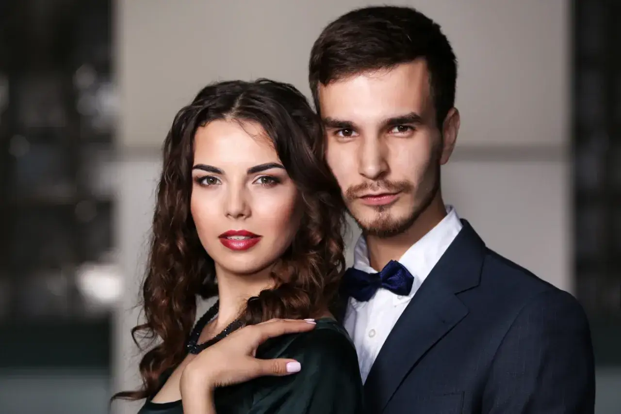Elegancka para, on w granatowym garniturze z muszką, ona w sukni z naszyjnikiem. Widać, jak kocha baran, gdy patrzy na nią z czułością.