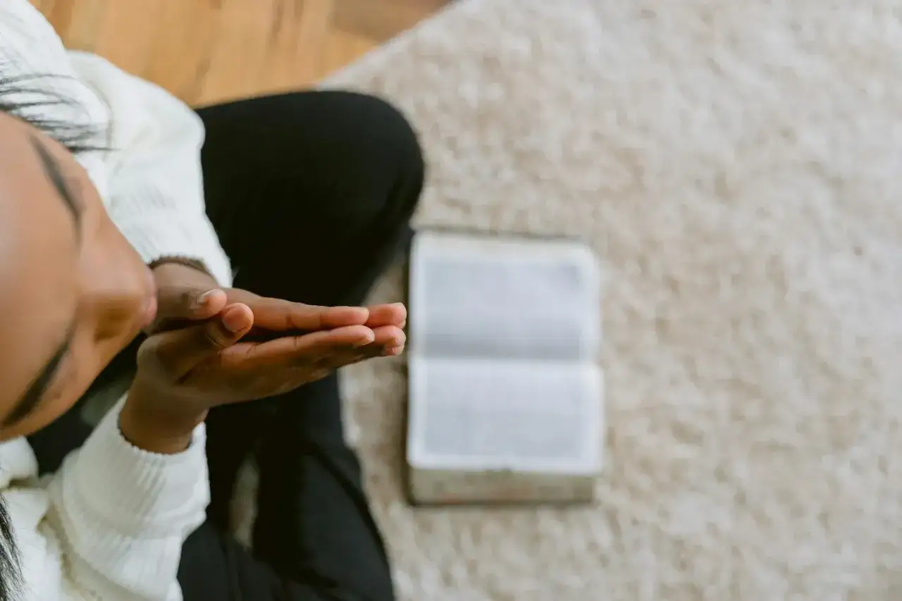 Osoba z zamkniętymi oczami składa dłonie do modlitwy. Obok leży otwarta księga, być może Biblia, symbolizująca krótką modlitwę o przebaczenie.