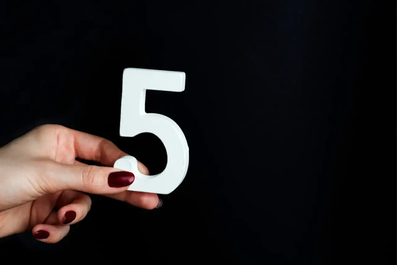 Dłoń z bordowymi paznokciami trzyma białą cyfrę 5, symbolizującą numerologiczną 5.